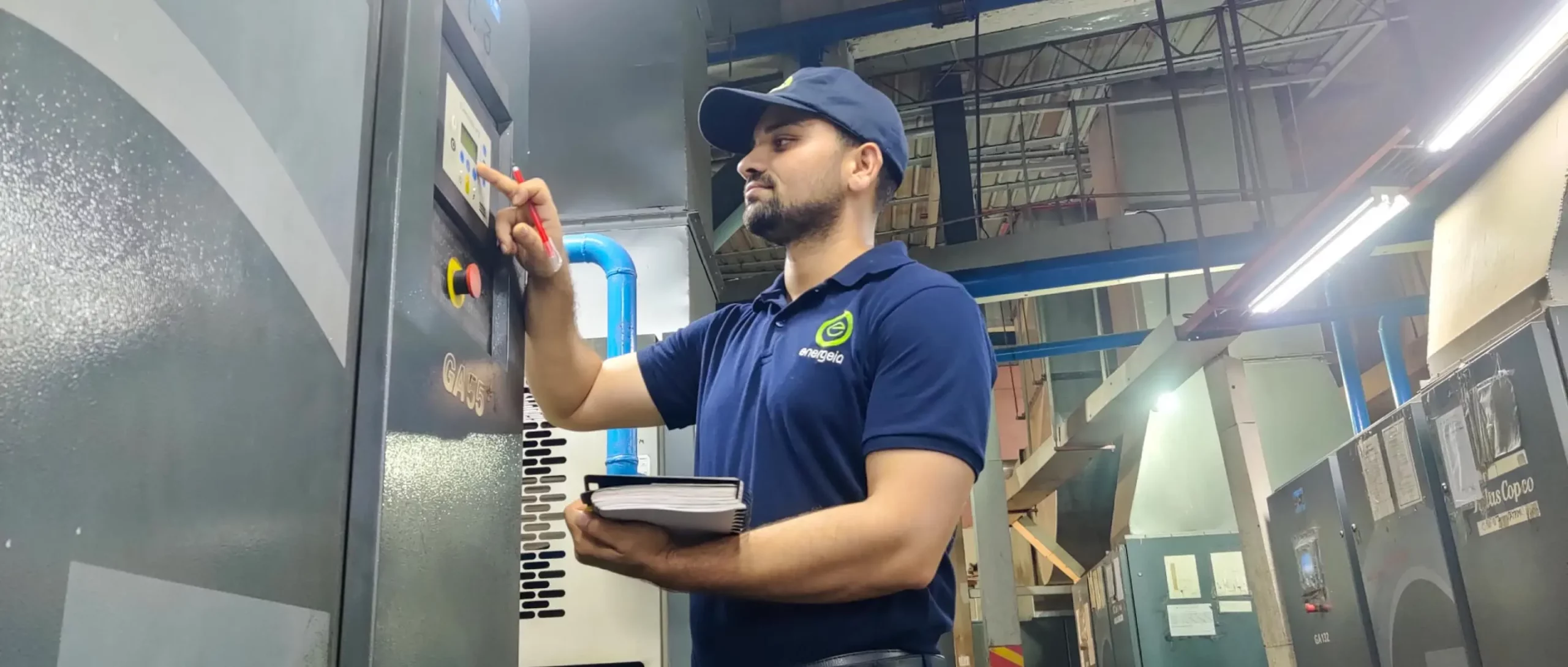 Technician in a blue Energeia uniform inspecting and adjusting a control panel on an industrial machine in a factory.