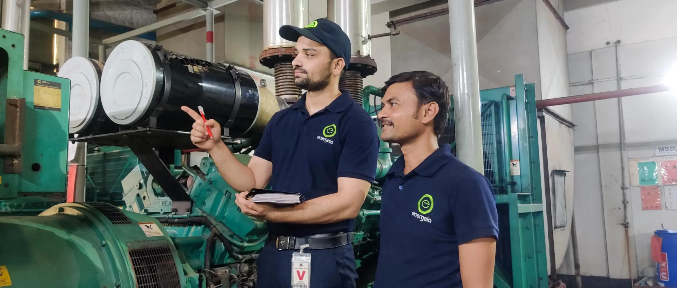 Two technicians in blue Energeia uniforms discussing an industrial system inside a factory, with one pointing towards equipment.