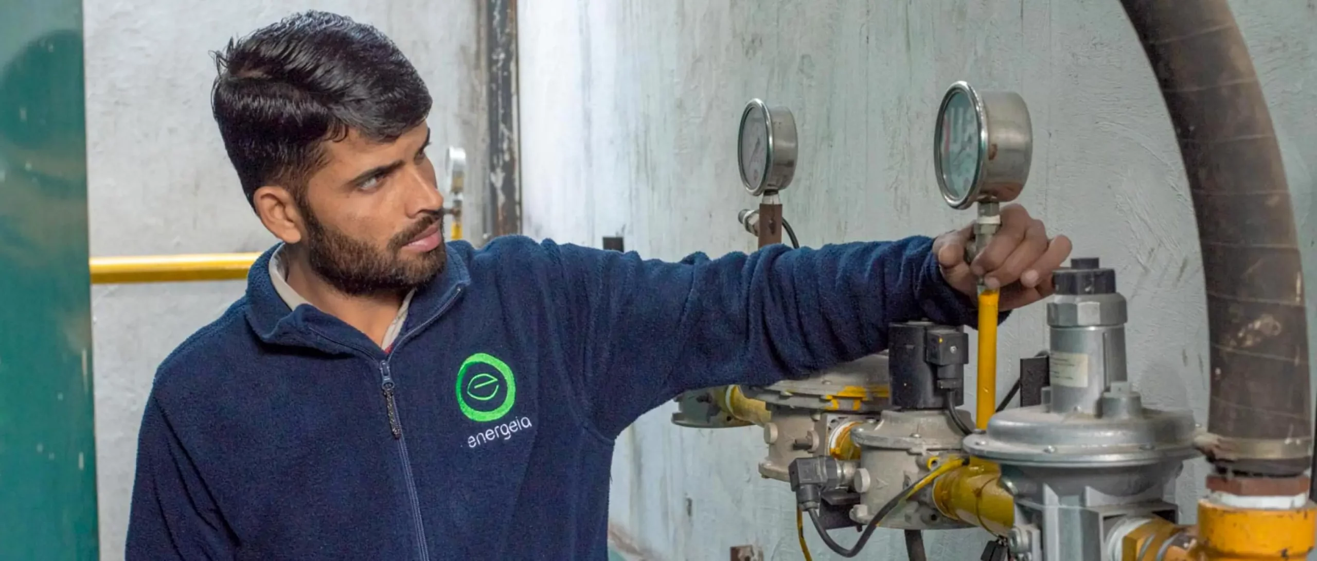 Technician in an Energeia fleece jacket inspecting a gas pressure gauge on industrial piping inside a facility.