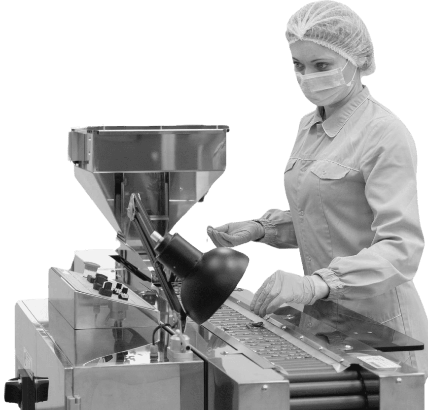 Black and white image of a female worker in a pharmaceutical or food production facility wearing a hairnet, face mask, and gloves while operating an industrial tablet or capsule filling machine.