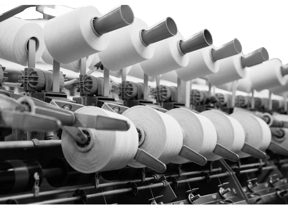 Black and white image of an industrial textile manufacturing process, showing multiple spools of white thread unwinding on an automated spinning machine, representing large-scale fabric production.