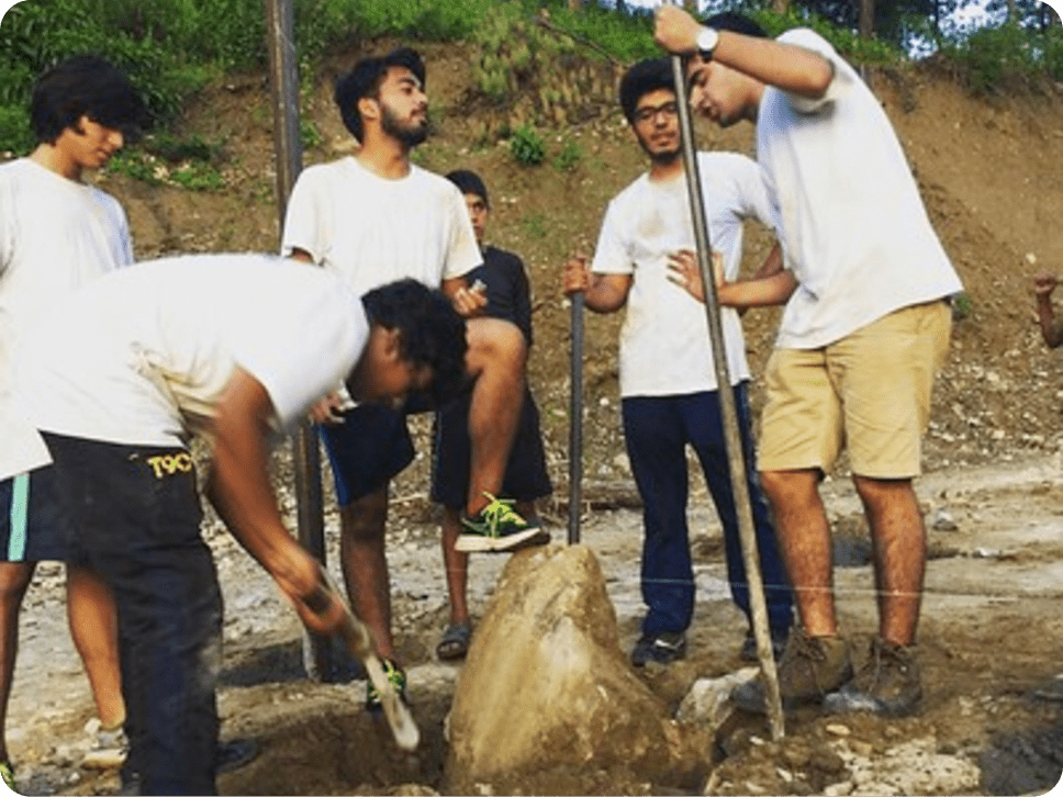 A group of young people wearing casual clothes and working together outdoors, using tools to dig and move rocks in a rugged, natural environment.
